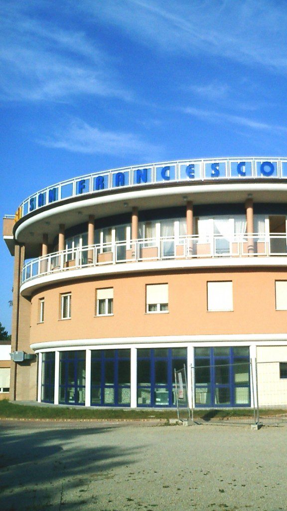 Edificio semicircolare arancione a due piani con finestroni e scritta blu "SAN FRANCESCO".