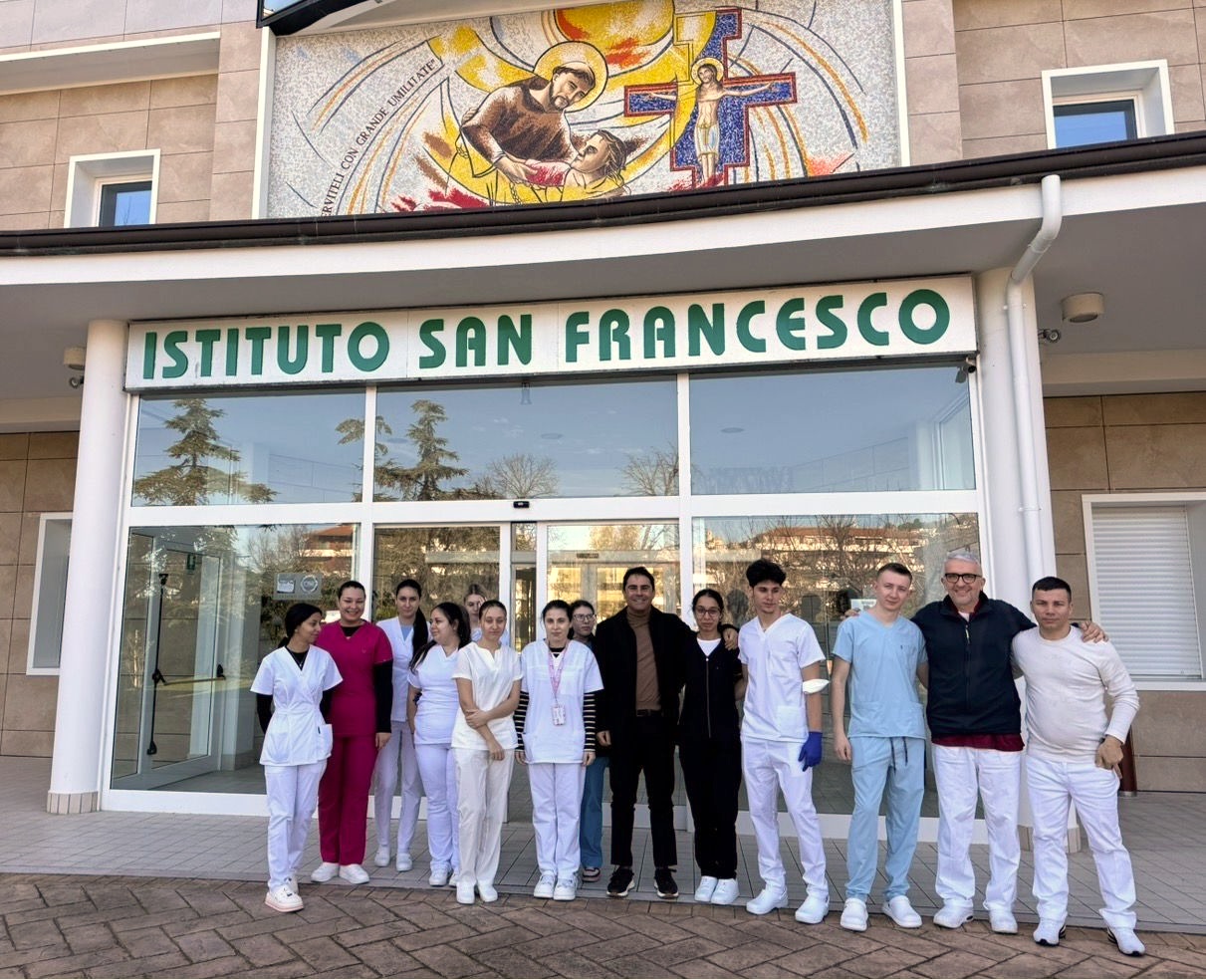 Gruppo di persone tra operatori sanitari davanti all'ingresso dell'Istituto San Francesco con murale religioso.